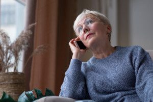 An elderly woman with short white hair is sitting by a window, holding a smartphone to her ear. She is wearing glasses and a gray sweater, and appears to be engaged in a phone conversation.